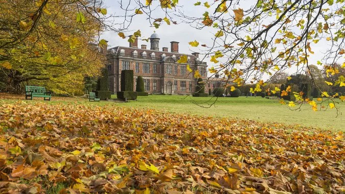 Ornate historic mansion in autumn surrounded by fallen leaves and colourful trees in a scenic park setting.